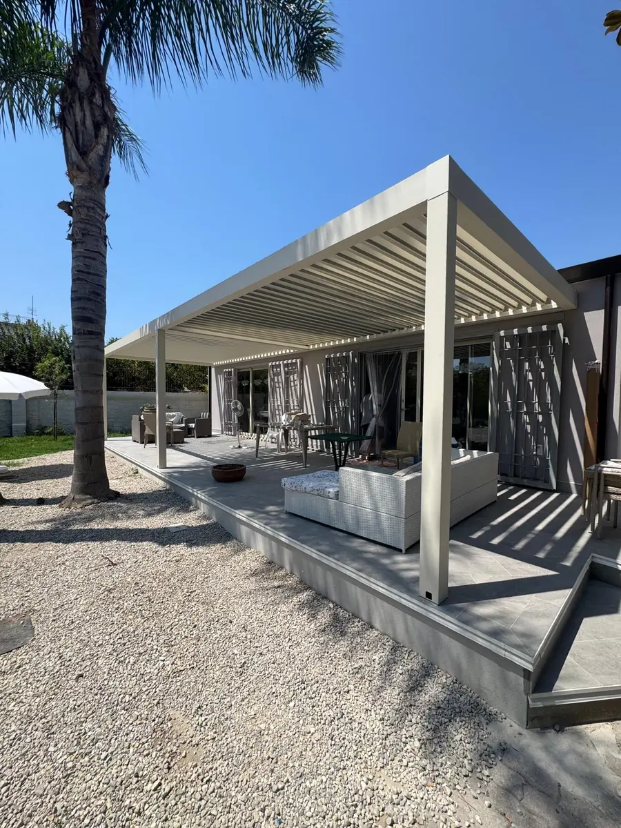 Pergola bioclimatica chiara in giardino, con lame aperte e zona soggiorno.