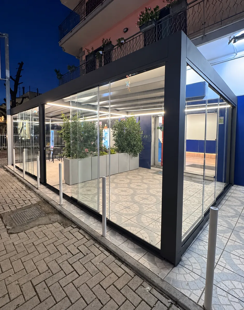 Dehors vetrato dai profili scuri con copertura illuminata da linee LED, visto di sera.