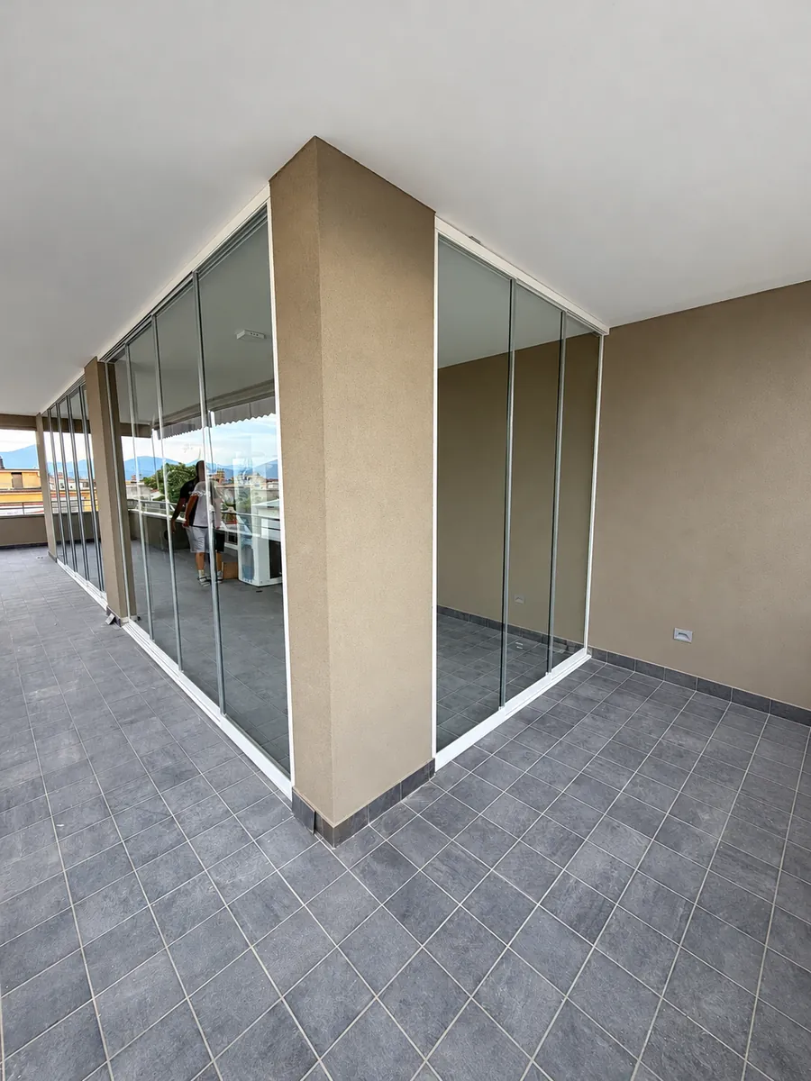Vetrate trasparenti lungo un balcone d’angolo, con pilastro centrale e pavimento grigio.