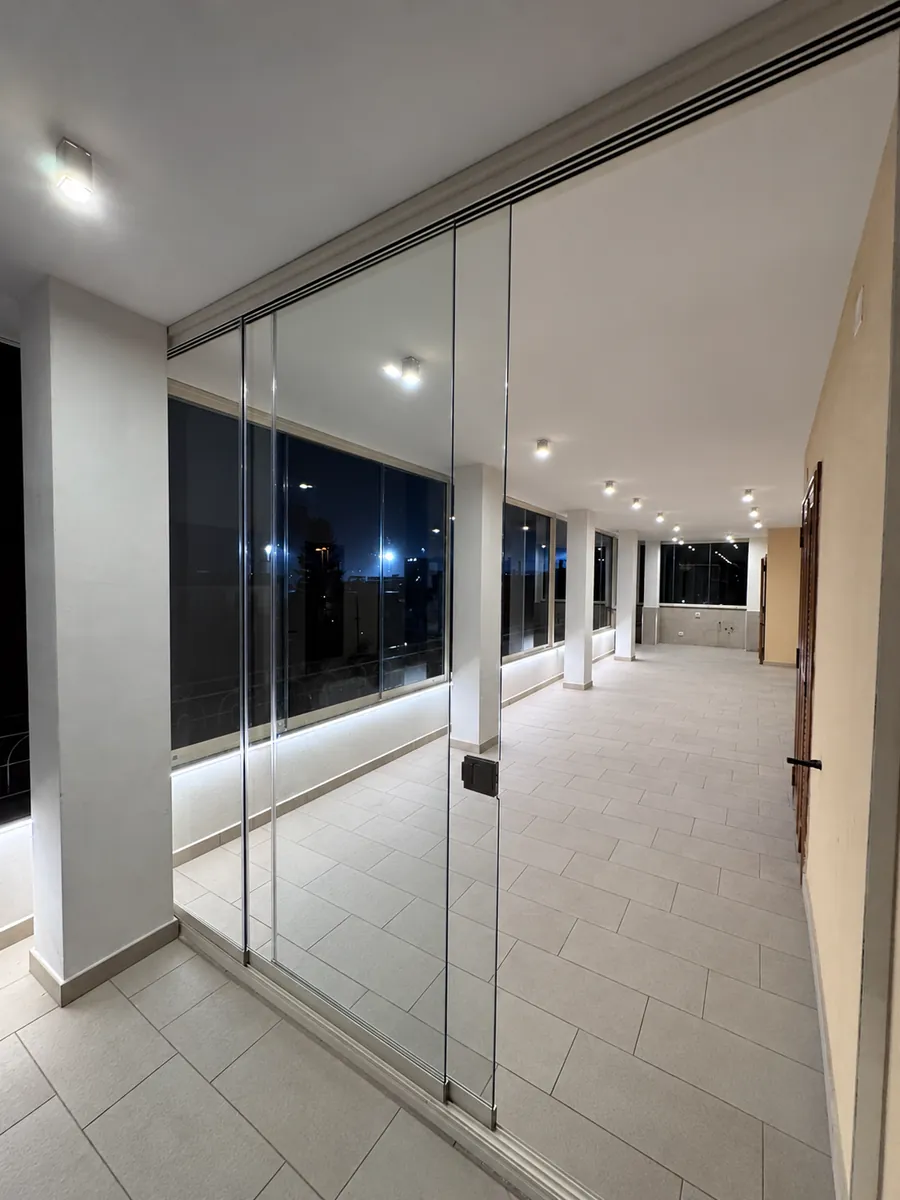Vetrate panoramiche lungo una loggia con pilastri, luci a soffitto e illuminazione sul parapetto.